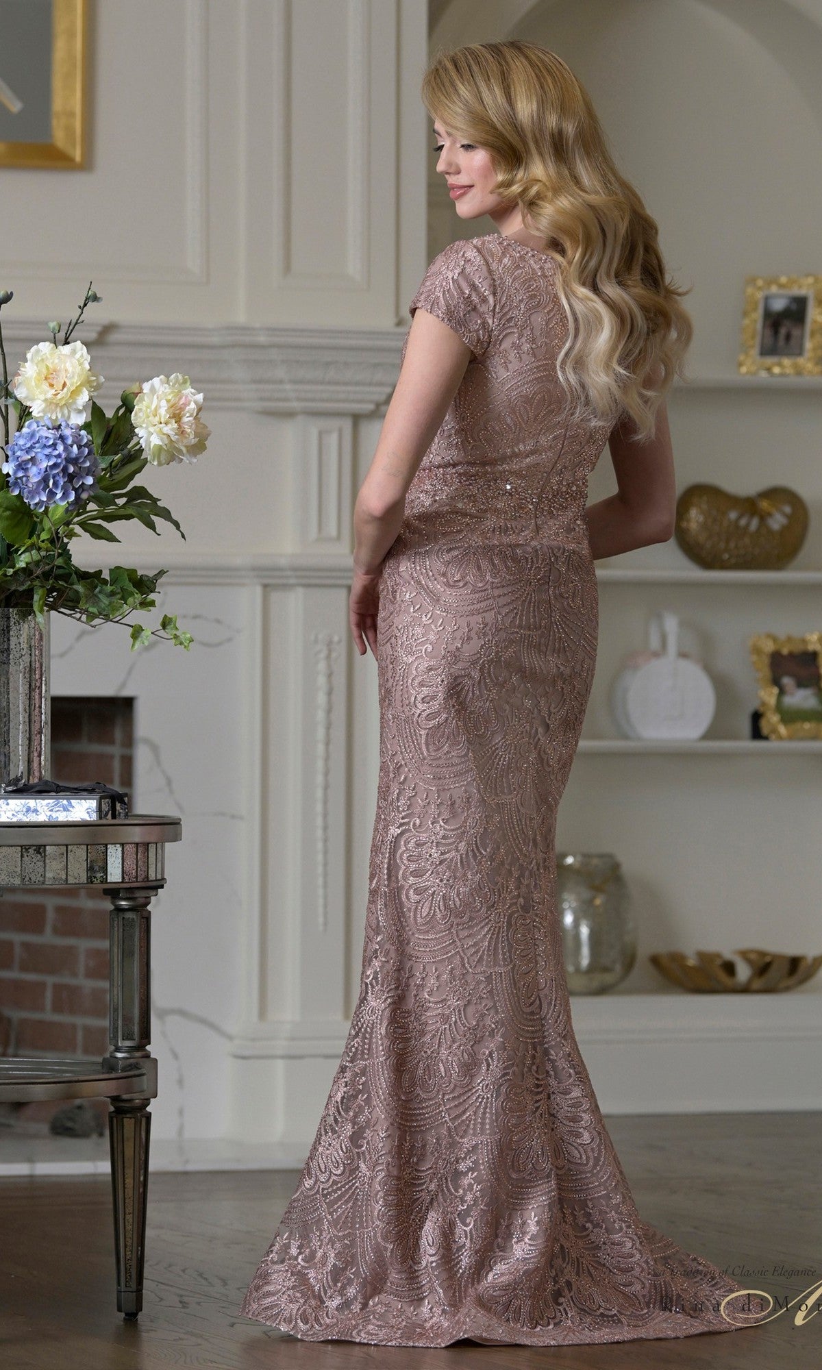 A woman with long blonde hair stands indoors, facing backward, wearing the fitted beaded Rina Di Montella RD3532 long formal dress with a mermaid silhouette. Shelves, a fireplace, and a table with flowers are in the background.